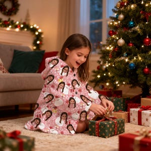 May include: A young person wearing a pink hooded blanket with a repeating pattern of faces, snowmen, and gift boxes. The person is opening a wrapped gift box near a decorated Christmas tree. Other wrapped gifts are scattered around.