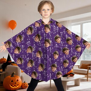 May include: A purple Halloween cape featuring a repeating pattern of white skeletons and a child's face. The cape has a hood and is worn by a child. Pumpkins and an orange balloon are in the background.