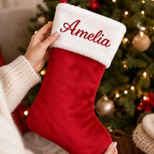 May include: A red velvet Christmas stocking featuring a white faux fur cuff at the top. The white cuff is embroidered with the name Amelia in a red cursive script. A small red fabric loop is attached to the top corner for hanging purposes.