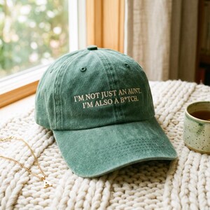 以下が含まれることがあります： 「I'M NOT JUST AN AUNT. I'M ALSO A B*TCH.」と刺繍されたグリーンのベースボールキャップ。帽子はクリーム色のニットブランケットの上に置かれています。金のネックレスと紅茶のマグカップも写っています。