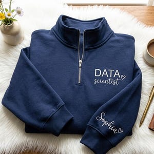 May include: A navy blue quarter-zip sweatshirt with the words "DATA scientist" and a heart graphic. The sleeve has the name "Sophia" embroidered. The sweatshirt is on a white fluffy surface.