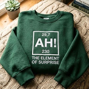 Pode incluir: Um moletom verde escuro com um gráfico branco que diz "AH!" e "THE ELEMENT OF SURPRISE". O moletom está dobrado sobre uma manta de malha bege perto de uma janela com uma pequena suculenta em vaso.
