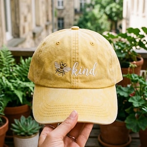 Op de afbeelding: Mosterdgele baseballcap met een gebogen klep. De cap heeft een geborduurde bij en het woord "kind" in witte letters. De cap is gemaakt van een zacht materiaal en heeft een klassiek zes-panelen ontwerp.