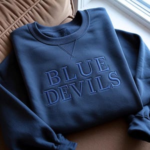 May include: A navy blue sweatshirt with the words "BLUE DEVILS" embroidered in a lighter shade of blue. The sweatshirt has a crew neck and ribbed cuffs. The garment is displayed on a brown surface.