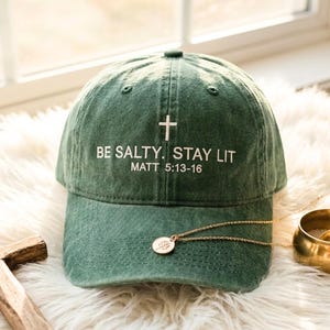 May include: A green baseball cap with the text "BE SALTY. STAY LIT" and a white cross. The text "MATT 5:13-16" is below the main text. A gold necklace and a small gold bowl are next to the cap.