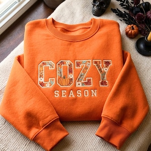 Pode incluir: Uma camisola laranja de gola redonda com o texto bordado COZY SEASON em letras de bloco. As letras estão preenchidas com um padrão detalhado de folhas de outono, pequenas flores brancas e abóboras laranja sobre um fundo creme.