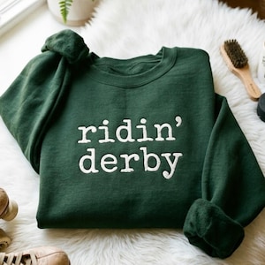 Puede incluir: Una sudadera verde oscuro con las palabras "ridin' derby" en letras blancas. La sudadera está doblada sobre una superficie blanca y esponjosa. Se ven patines en la esquina inferior izquierda.