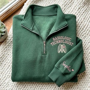 Könnte beinhalten: Dunkelgrünes Sweatshirt mit Viertelreißverschluss. Es zeigt die Worte "RADIOLOGIC TECHNOLOGIST" und ein rosafarbenes Rippendesign. Der Ärmel hat den Namen "Sophie" in rosa Schrift mit einem Herz.