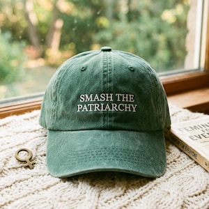 Puede incluir: Gorra de béisbol verde con la frase "SMASH THE PATRIARCHY" bordada en blanco. La gorra está sobre una superficie de punto color crema, con un amuleto dorado con el símbolo femenino y un libro titulado "FEMINISM FOR EVERYONE".