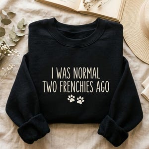 K&ouml;nnte beinhalten: Schwarzes Sweatshirt mit dem wei&szlig;en Text "I WAS NORMAL TWO FRENCHIES AGO" und zwei wei&szlig;en Pfotenabdr&uuml;cken. Das Kleidungsst&uuml;ck wird auf einer neutralen Oberfl&auml;che pr&auml;sentiert, mit einem Buch und einem Hut im Hintergrund.