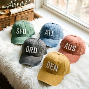 May include: Five baseball caps in various colors: green, blue, gray, rust, and yellow. Each cap has a different three-letter city abbreviation in white block letters: SFO, ATL, ORD, AUS, and DEN. The caps are displayed on a fluffy white surface.