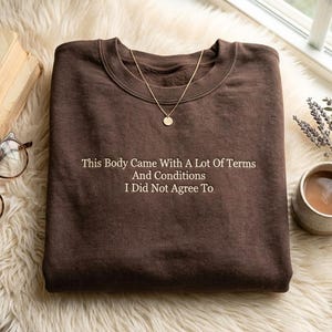 Op de afbeelding: Een bruine sweatshirt met de tekst "This Body Came With A Lot Of Terms And Conditions I Did Not Agree To" in het wit. Een gouden ketting met een hanger ligt op de sweatshirt. De sweatshirt ligt op een zacht, wit oppervlak.