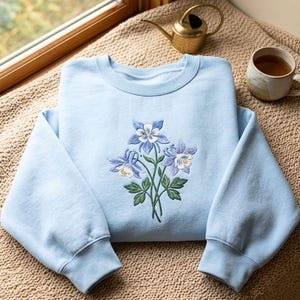 May include: A light blue crewneck sweatshirt featuring an embroidered design of three columbine flowers with blue and white petals and green leaves centered on the chest. The garment has long sleeves with ribbed cuffs and a ribbed neckline.
