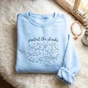 Könnte beinhalten: Hellblaues Sweatshirt mit dem Schriftzug "protect the sharks" in dunkelblauer Schrift. Das Design zeigt verschiedene Hai-Illustrationen in Dunkelblau, darunter einen Hammerhai und einen Walhai. Das Sweatshirt ist aus weichem Material.