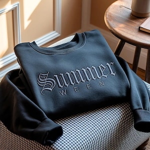 May include: A dark gray sweatshirt with the word "Summer" embroidered in a gothic font. The sweatshirt is folded on a chair with a black and white checkered pattern. A wooden side table and a mug are in the background.
