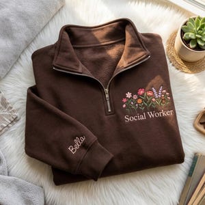Könnte beinhalten: Ein braunes Sweatshirt mit Viertelreißverschluss, bestickt mit floralem Design und dem Text "Social Worker". Am linken Ärmelbund ist der Name "Bella" eingestickt. Das Sweatshirt ist aus weichem Material.