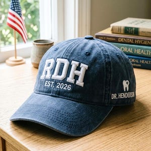 Könnte beinhalten: Marineblaue Baseballkappe mit weiß gesticktem Text "RDH EST. 2026" und "DR. HENDERSON" mit einer Zahngrafik. Die Kappe liegt auf einer Holzoberfläche, mit einer amerikanischen Flagge im Hintergrund.