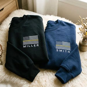 Puede incluir: Dos sudaderas de cuello redondo, una negra y otra azul marino, cada una con un pequeño diseño de bandera estadounidense con una línea amarilla y un nombre debajo. La sudadera negra tiene impreso "MILLER", mientras que la azul marino dice "SMITH".