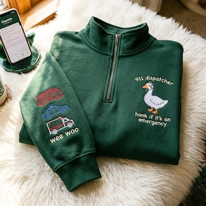 Könnte beinhalten: Ein dunkelgrünes Sweatshirt mit Viertelreißverschluss und gestickten Notdienstfahrzeugen auf dem linken Ärmel. Die rechte Seite zeigt eine Gans mit Headset und dem Text "911 dispatcher, honk if it's an emergency". Das Sweatshirt liegt auf einer weißen, flauschigen Oberfläche.