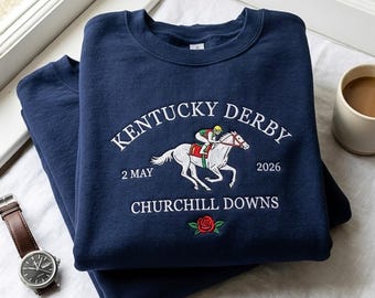 May include: Navy blue sweatshirt featuring embroidered "Kentucky Derby" text, a horse and jockey graphic, and the date "2 May 2026". A red rose is embroidered below the text. A watch and a cup of coffee are in the background.