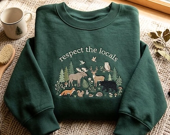 May include: A dark forest green crewneck sweatshirt featuring an embroidered graphic of various woodland animals including a moose, deer, bear, fox, and owl, with the text "respect the locals" arched above the scene in white lettering.