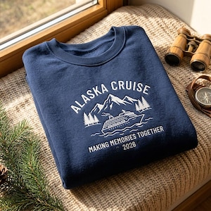 Op de afbeelding: Een marineblauw sweatshirt met ronde hals en een witte grafische print van een cruiseschip, een bergketen en dennenbomen. De tekst luidt ALASKA CRUISE, MAKING MEMORIES TOGETHER en 2026, gecentreerd op de borst van het kledingstuk.