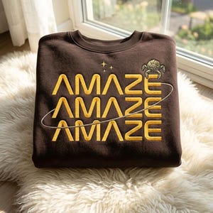 Könnte beinhalten: Ein braunes Sweatshirt mit dem Wort "AMAZE" in Gold, dreimal wiederholt. Eine kleine Grafik einer Kreatur und Sterne befinden sich über dem Text. Eine weiße Linie umkreist den Text. Das Sweatshirt liegt auf einer weißen, flauschigen Oberfläche.