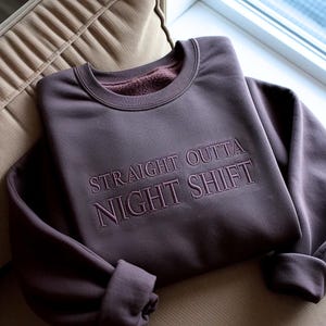 May include: A mauve-colored sweatshirt with the embroidered text "STRAIGHT OUTTA NIGHT SHIFT" in a darker shade. The sweatshirt is folded on a beige cushion, with the sleeves rolled up.