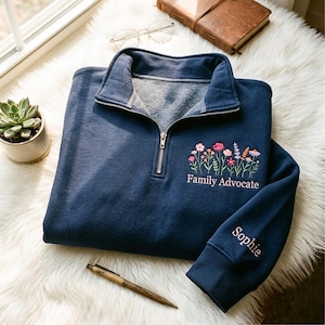 Könnte beinhalten: Ein marineblaues Sweatshirt mit einem Viertel-Reißverschluss-Ausschnitt. Es hat ein gesticktes Blumendesign und die Worte "Family Advocate". Der Ärmel ist mit dem Namen "Sophie" bestickt.