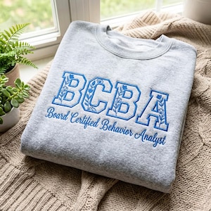 Könnte beinhalten: Ein graues Sweatshirt mit dem blau gestickten Text "BCBA", mit floralen Details in den Buchstaben. Darunter ist der Text "Board Certified Behavior Analyst" ebenfalls blau gestickt. Das Sweatshirt liegt gefaltet auf einer beigen Strickdecke.