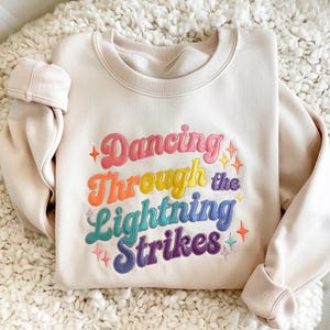 May include: Cream-colored sweatshirt with the embroidered text "Dancing Through the Lightning Strikes" in rainbow colors. The text is in a retro font with star accents. The sweatshirt has a crew neck and ribbed cuffs.