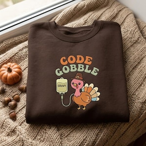 Può includere: Una felpa girocollo marrone scuro con la grafica di un tacchino che indossa un cappello da pellegrino. Il tacchino è collegato a una sacca per flebo con l'etichetta GRAVY. Sopra il tacchino, il testo recita CODE GOBBLE in font retro arancione.
