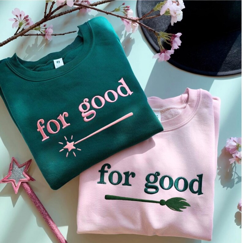 K&ouml;nnte beinhalten: Zwei Sweatshirts, eines in Smaragdgr&uuml;n und eines in Hellrosa, werden gezeigt. Das gr&uuml;ne Sweatshirt hat die Worte "for good" in Rosa mit einem Zauberstab-Design gestickt. Das rosa Sweatshirt hat denselben Text in Gr&uuml;n mit einem Besen-Design.