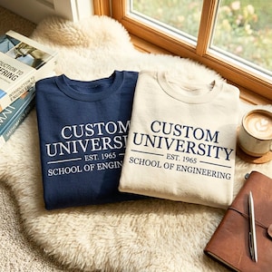 Könnte beinhalten: Zwei Sweatshirts, eines marineblau und eines cremefarben, mit dem Text "CUSTOM UNIVERSITY EST. 1965 SCHOOL OF ENGINEERING" in Weiß aufgedruckt. Die Sweatshirts liegen auf einem flauschigen weißen Teppich in der Nähe eines Fensters.