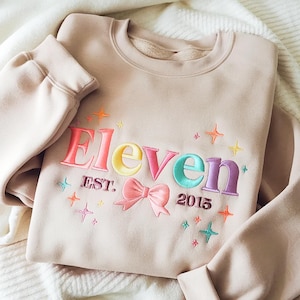 May include: A beige sweatshirt with the word "Eleven" embroidered in colorful letters, a pink bow, and the year "2015". The design includes star accents in various colors. The sweatshirt is folded on a white surface.