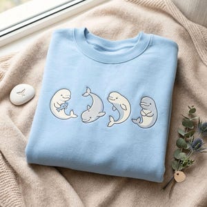 May include: A light blue crewneck sweatshirt featuring an embroidered graphic of four white and grey beluga whales in various poses, including one whale holding a small blue fish, arranged horizontally across the centre of the garment.