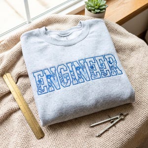 Puede incluir: Sudadera gris claro con la palabra "ENGINEER" bordada en azul, con detalles florales dentro de las letras. Se ven una regla de latón y un compás.