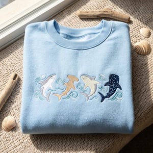 Puede incluir: Una sudadera de cuello redondo en color azul claro con un diseño bordado de cuatro tiburones diferentes nadando entre olas estilizadas de color verde azulado. Los tiburones incluyen un tiburón gris, un tiburón martillo beige, uno crema y un tiburón ballena azul oscuro.