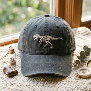 Op de afbeelding: Een antracietgrijze baseballpet met een geborduurd wit T-Rex dinosaurusskelet op het voorpaneel. De pet heeft een gestructureerde kroon, een gebogen klep en gestikte oogjes voor ventilatie in een klassiek zes-panelen ontwerp.