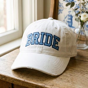 Puede incluir: Gorra de béisbol blanca con la palabra "BRIDE" bordada en azul. La gorra tiene una visera curva y un aspecto desgastado. Descansa sobre una superficie de madera.