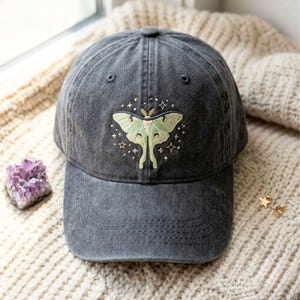 May include: A charcoal gray baseball cap featuring an embroidered light green luna moth centered on the front panel. The moth is surrounded by small stitched stars and sparkles in white and gold thread, with a curved brim and six-panel design.