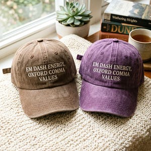 Può includere: Vengono mostrati due cappellini da baseball, uno marrone e uno viola. Ogni cappellino presenta il testo bianco "EM DASH ENERGY, OXFORD COMMA VALUES". I cappellini sono su una superficie strutturata vicino a una finestra, con libri e una tazza di tè sullo sfondo.