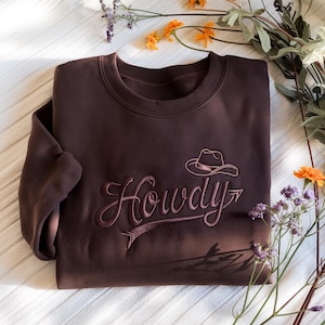 May include: A dark brown sweatshirt with the word "Howdy" embroidered in a cursive font, along with a cowboy hat graphic. The design is in a lighter brown color. The sweatshirt is surrounded by flowers.