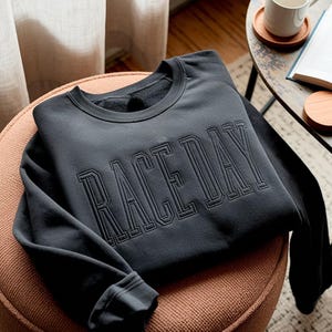 Może przedstawiać: Czarna bluza z okrągłym dekoltem z napisem "RACEDAY" wyhaftowanym na piersi w stylu dużych liter. Bluza jest starannie złożona na okrągłym, tapicerowanym stołku. Rękawy są podwinięte, a materiał wydaje się miękki.