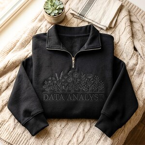 Puede incluir: Una sudadera negra con cuello con cremallera y diseño floral bordado. El texto "DATA ANALYST" está bordado debajo del diseño floral. La sudadera está doblada sobre una manta de punto beige.