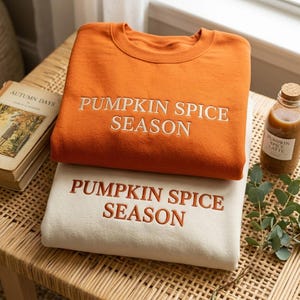 Op de afbeelding: Een roestoranje sweater met ronde hals en witte geborduurde tekst PUMPKIN SPICE SEASON, gestapeld op een crèmekleurige sweater met ronde hals en roestoranje geborduurde tekst PUMPKIN SPICE SEASON.