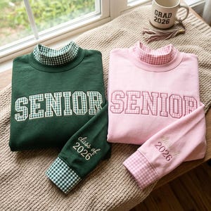 Puede incluir: Dos sudaderas de manga larga, una verde oscuro y otra rosa claro, con cuello alto y puños de patrón de cuadros vichy. La palabra SENIOR está bordada en cuadros vichy en el pecho, y la manga muestra el texto class of 2026 en cursiva.