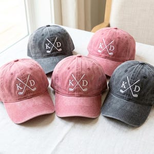 Può includere: Sono mostrati cinque cappellini da golf in tonalità rosa e grigio. Ogni cappellino presenta le lettere ricamate "KXD" con mazze da golf incrociate. I cappellini sono realizzati in un tessuto morbido e lavato e hanno un classico design da baseball.