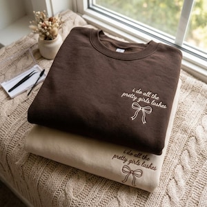 Könnte beinhalten: Ein dunkelbraunes Sweatshirt und ein beiges Sweatshirt liegen übereinander. Beide zeigen den gestickten Text "i do all the pretty girls lashes" in Schreibschrift, ergänzt durch ein kleines Linien-Symbol einer Schleife unter dem Text.