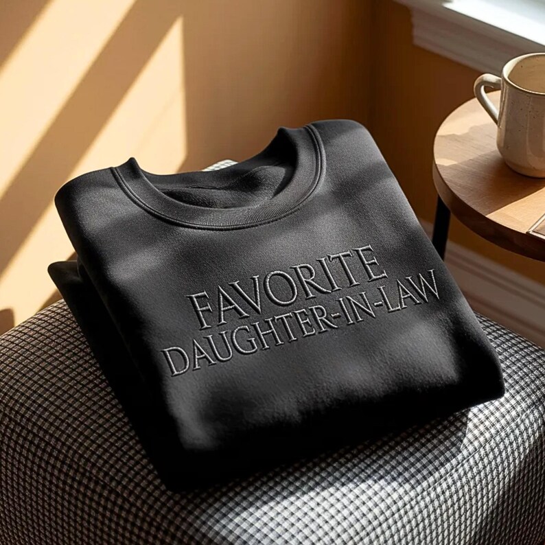 May include: A black sweatshirt with the embroidered text "FAVORITE DAUGHTER-IN-LAW" in a light grey color. The sweatshirt is folded and resting on a patterned cushion. A beige mug sits on a small wooden table in the background.